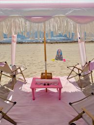Sunny oceanfront beach setup under a pink fringed umbrella: pink mat and low pink table with heart-shaped sunglasses, two folding beach chairs on the sand, colorful boogie board and sandals near the shoreline.