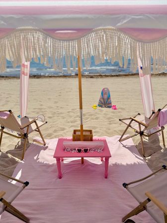 Sunny oceanfront beach setup under a pink fringed umbrella: pink mat and low pink table with heart-shaped sunglasses, two folding beach chairs on the sand, colorful boogie board and sandals near the shoreline.