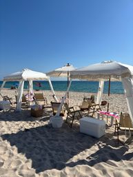 Sunny sandy beach with white fringed canopies, wooden lounge chairs, coolers and beach gear set up along the turquoise ocean under a clear blue sky — vacation-ready seaside scene.