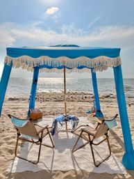 Sunlit coastal scene with a blue fringed beach canopy shading two folding chairs on a white blanket, picnic basket and nautical netting on sandy shore overlooking sparkling ocean waves and bright sky.