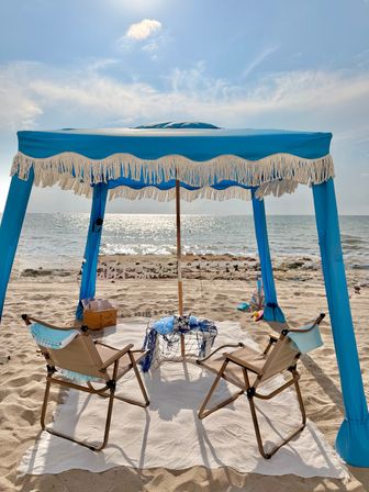 Sunlit coastal scene with a blue fringed beach canopy shading two folding chairs on a white blanket, picnic basket and nautical netting on sandy shore overlooking sparkling ocean waves and bright sky.