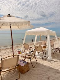Beach cabana and fringed white umbrellas shading folding chairs on a sandy shore, calm turquoise ocean with a few swimmers and a textured cloudy sky
