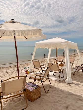 Beach cabana and fringed white umbrellas shading folding chairs on a sandy shore, calm turquoise ocean with a few swimmers and a textured cloudy sky