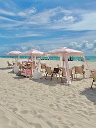 Playful pink-and-white striped cabanas and umbrellas with folding beach chairs on white sand, turquoise ocean and blue sky with scattered clouds on a sunny beach day.