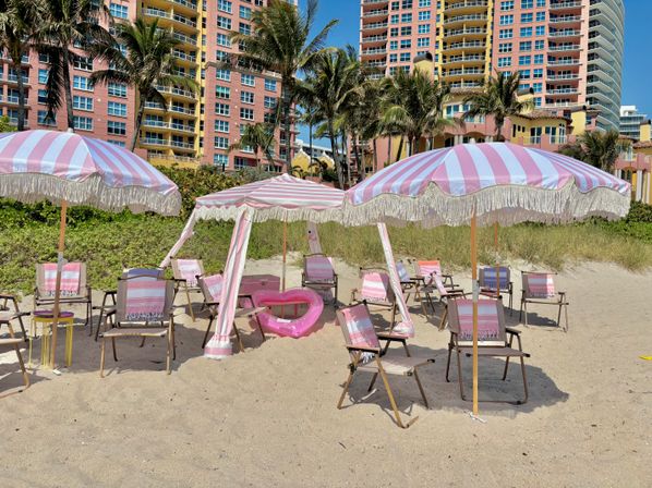 Sunny beachfront scene with pink-and-white striped fringe umbrellas, matching chairs and towels, a pink inflatable float on sand, palm trees and pastel high-rise condos in the background.