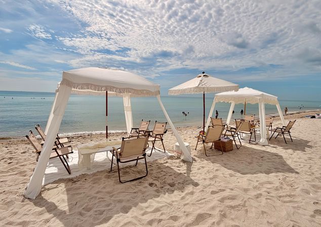 Sandy beach with white fringe-canopy cabanas and umbrellas, folding lounge chairs on the shore facing calm turquoise ocean under a dramatic partly-cloud sky — serene tropical seaside scene.