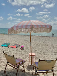 Sunny seaside scene with two canvas beach chairs and a pink-and-white striped fringe umbrella over a small table on soft sand, turquoise ocean and clouds in the background, cornhole boards and beach toys nearby, and a distant silhouetted figure by the water.