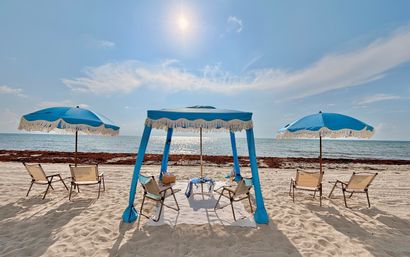 Sunny sandy beach with sparkling ocean, a blue fringed cabana flanked by two blue umbrellas and lounge chairs ready for relaxing.