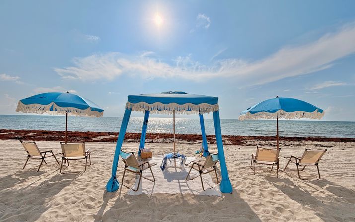 Sunny sandy beach with sparkling ocean, a blue fringed cabana flanked by two blue umbrellas and lounge chairs ready for relaxing.