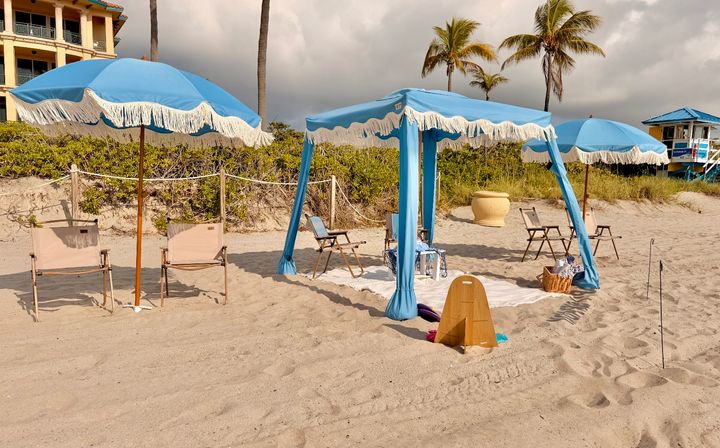 Coastal beach scene with turquoise fringed umbrellas and matching canopy over folding chairs and a picnic setup on sandy shore, palm trees and a lifeguard hut under a cloudy sky.