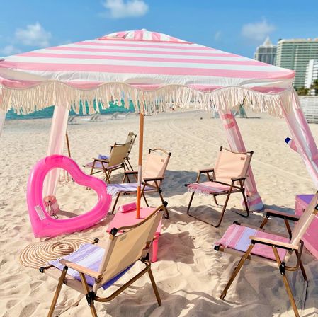 Pink-and-white striped fringe beach umbrella shading folding chairs and a bright pink heart-shaped float on a sunny sandy beach with turquoise ocean