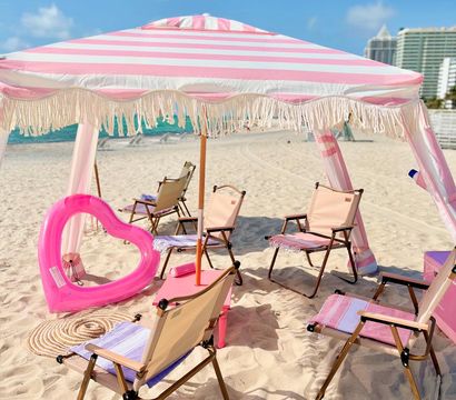 Pink-and-white striped fringe beach umbrella shading folding chairs and a bright pink heart-shaped float on a sunny sandy beach with turquoise ocean