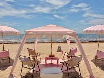 Pink-and-white striped fringe canopy and matching umbrellas shading folding chairs and a small pink table on a sunny sandy beach, calm blue ocean and wispy clouds on the horizon.