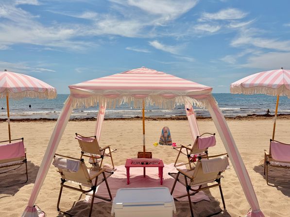 Pink-and-white striped fringe canopy and matching umbrellas shading folding chairs and a small pink table on a sunny sandy beach, calm blue ocean and wispy clouds on the horizon.