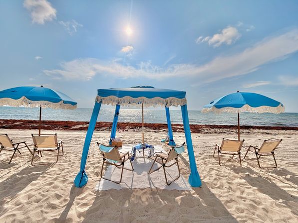 Bright sunny beach with a blue fringed canopy and matching umbrellas shading folding lounge chairs on white sand, calm sparkling ocean and clear blue sky.