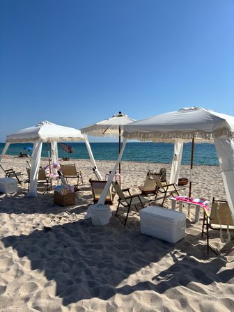 Sunny beach scene with white fringe cabanas and umbrellas, wooden lounge chairs and coolers on sandy shore facing turquoise ocean under a clear blue sky.