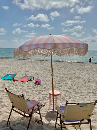 Sunny sandy beach with turquoise ocean and blue sky; pastel pink-and-white striped fringe umbrella shades two canvas lounge chairs, cornhole boards and beach toys nearby.