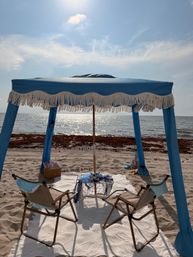 Sunlit seaside setup on white sand: blue fringed canopy with two beach chairs and a small nautical-decor table facing the sparkling ocean
