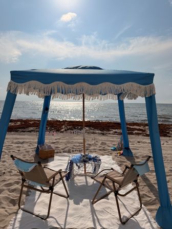Sunlit seaside setup on white sand: blue fringed canopy with two beach chairs and a small nautical-decor table facing the sparkling ocean