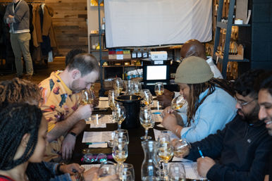 Group wine-tasting class at a long table — people sampling white wines, sniffing glasses and jotting tasting notes in a cozy indoor space