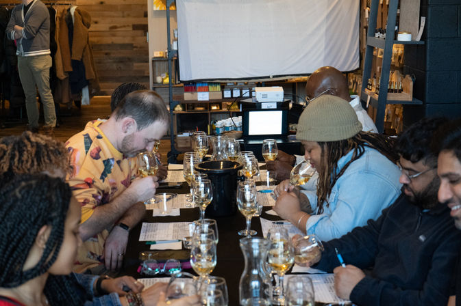 Group wine-tasting class at a long table — people sampling white wines, sniffing glasses and jotting tasting notes in a cozy indoor space