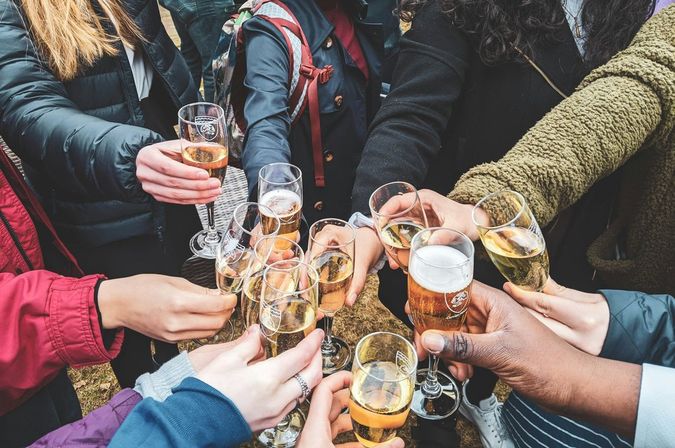 Many hands in jackets clinking champagne flutes and a beer glass in an outdoor celebration, a bubbly toast among a group.