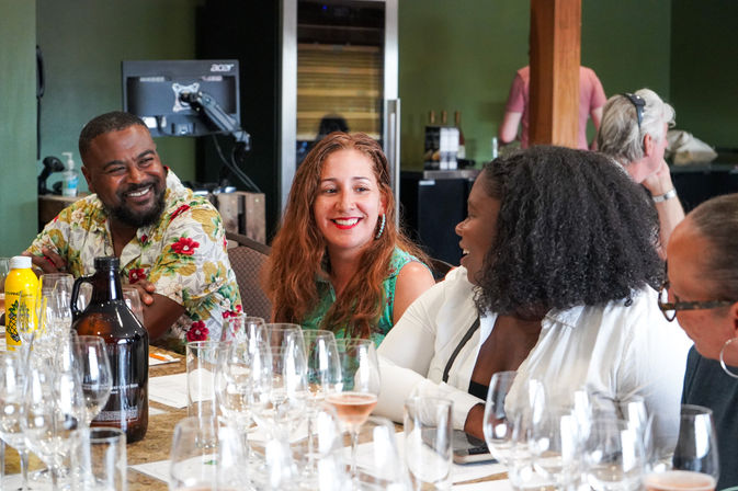 Smiling group laughing and chatting during a lively wine tasting in a cozy tasting room, surrounded by wine glasses, a growler, and tasting notes.