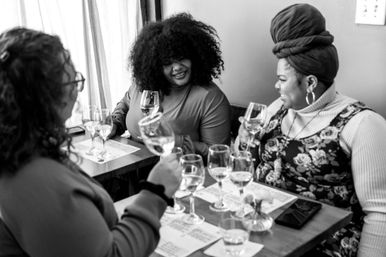 Three friends enjoying a wine tasting at a cozy wine bar, smiling around a table with multiple tasting glasses and notes.