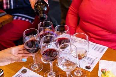 Red wine being poured into a tasting flight of wine glasses on a wooden table with tasting menus nearby.