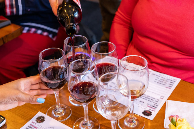 Red wine being poured into a tasting flight of wine glasses on a wooden table with tasting menus nearby.