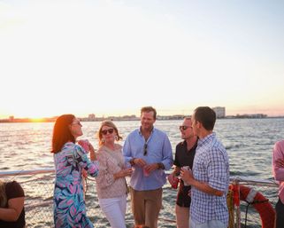 Group of friends chatting and holding drinks on a boat deck at sunset, coastal skyline and calm harbor water in the background.