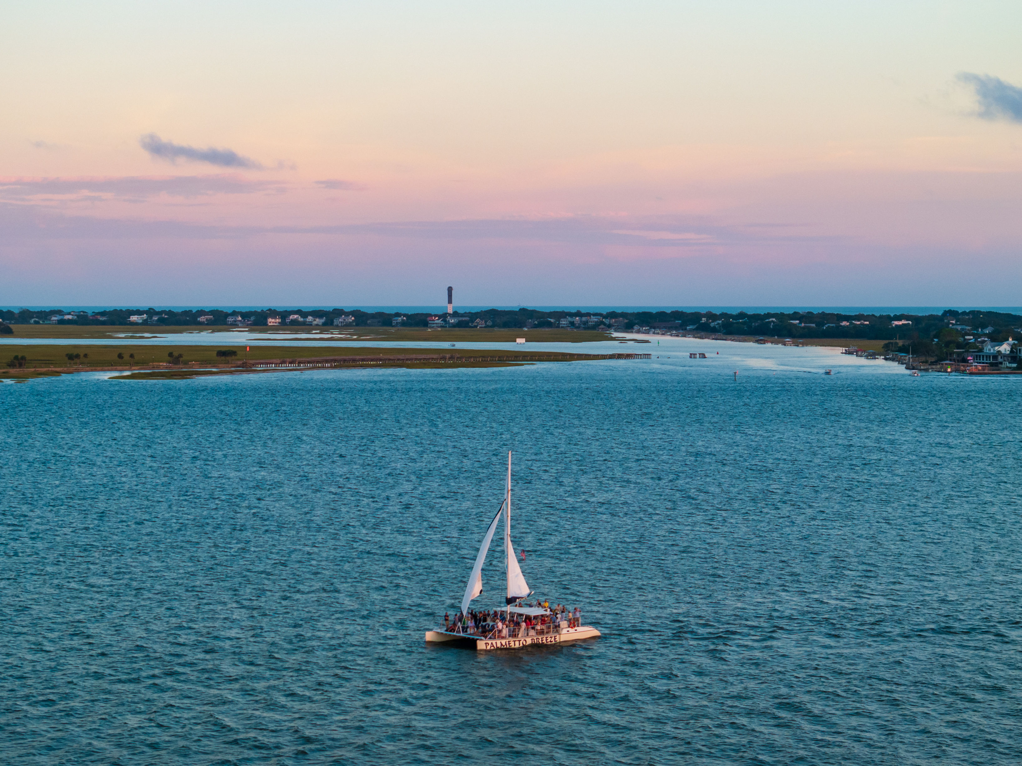 Catamaran of passengers sailing on a blue coastal inlet at sunset with a pastel pink sky, marshy shoreline and a distant lighthouse on the horizon.
