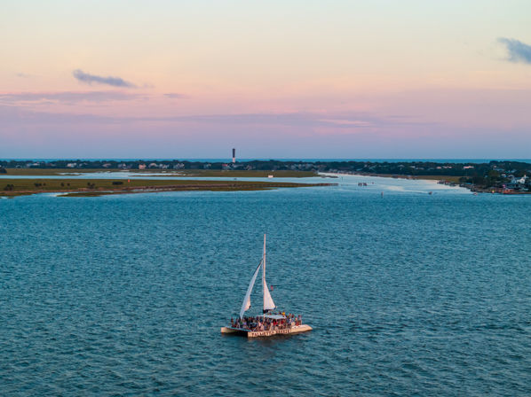 Catamaran of passengers sailing on a blue coastal inlet at sunset with a pastel pink sky, marshy shoreline and a distant lighthouse on the horizon.