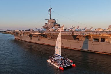Passenger catamaran sailing past a docked aircraft carrier lined with naval planes during golden-hour, warm sunset light on calm harbor waters.