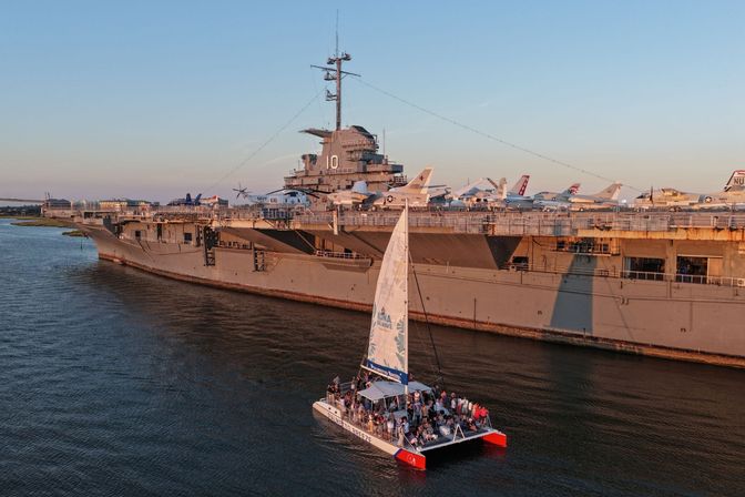 Passenger catamaran sailing past a docked aircraft carrier lined with naval planes during golden-hour, warm sunset light on calm harbor waters.