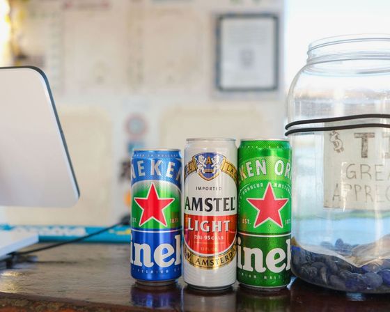 Three beer cans—Amstel Light and two Heineken—lined up on a bar counter next to a tip jar and a point-of-sale screen