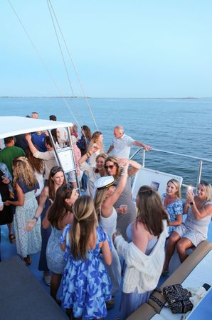 Group of friends enjoying a lively yacht deck party with ocean views — women in summer dresses dancing, laughing and taking photos during a coastal evening boat cruise.
