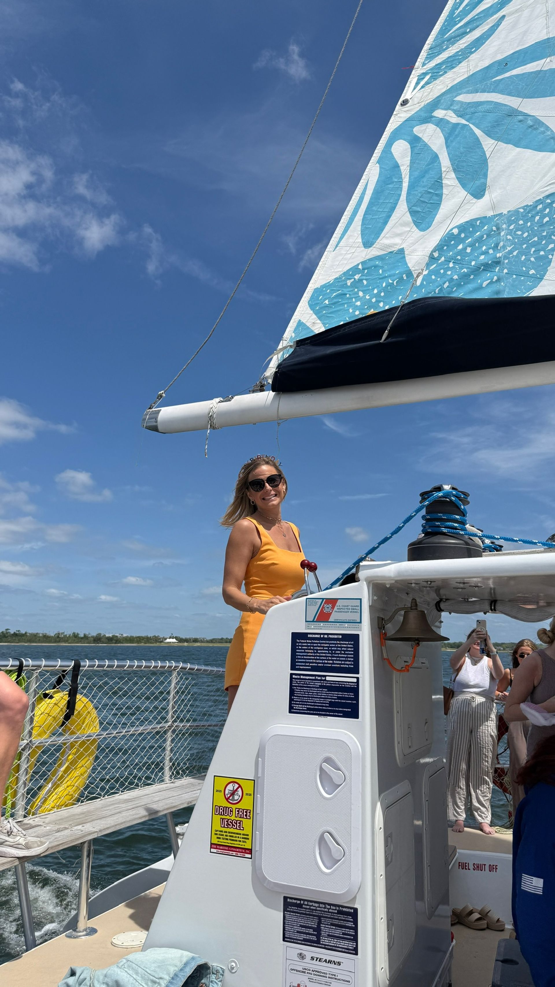 Woman in a yellow sundress and sunglasses at the helm of a sailboat on a sunny coastal ocean day, blue patterned sail overhead with other passengers and a life ring visible.