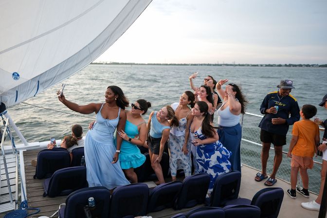 Smiling group of friends in blue summer dresses taking a selfie on a sailboat during a coastal sunset cruise with ocean in the background