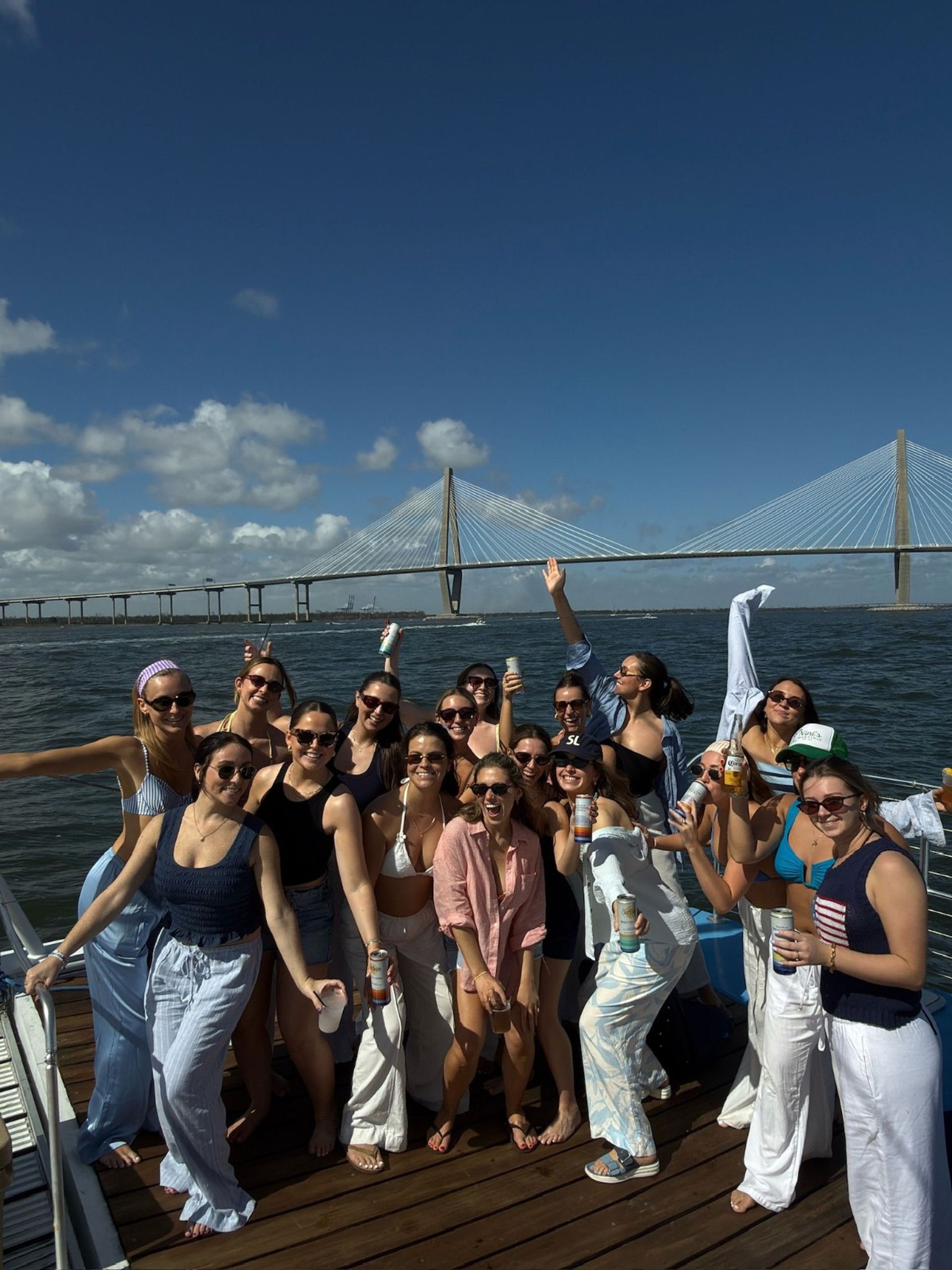 Group of friends on a boat holding drinks and smiling with a cable-stayed bridge, sunny blue sky, and harbor in the background