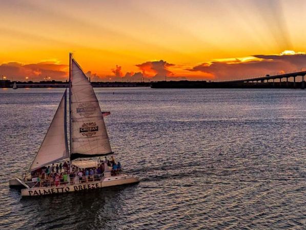 Catamaran crowded with passengers on a sunset sail across a calm coastal bay, warm orange sky and a long bridge on the horizon