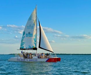 Sunlit catamaran with a tropical-patterned sail and passengers on deck cruising on calm blue coastal waters under a clear sky