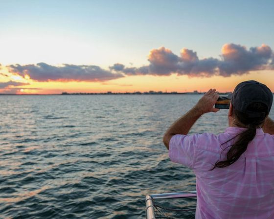 Person in pink shirt and cap on a boat photographing a vibrant coastal sunset over rippling ocean waters with distant shoreline and dramatic clouds.