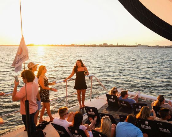 Group of people socializing and laughing on a boat deck at sunset, enjoying a warm golden harbor cruise with a distant city skyline and calm water.