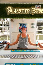Smiling crew member in a blue cap and gray tank top standing behind a boat-side service counter at a sunny coastal marina, with a sunglasses display and pier in the background.