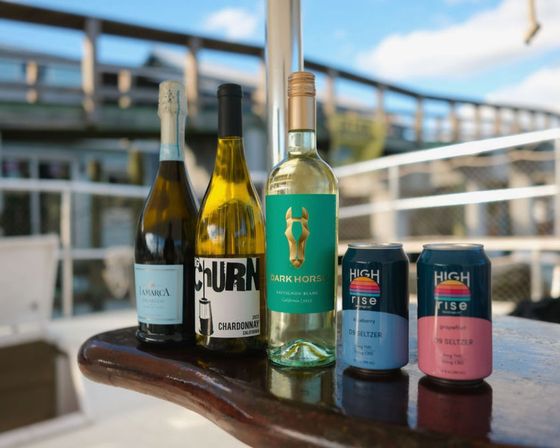 Three white wine bottles and two colorful hard seltzer cans lined up on a glossy wooden boat-deck railing with a sunny marina pier and blue sky in the background.