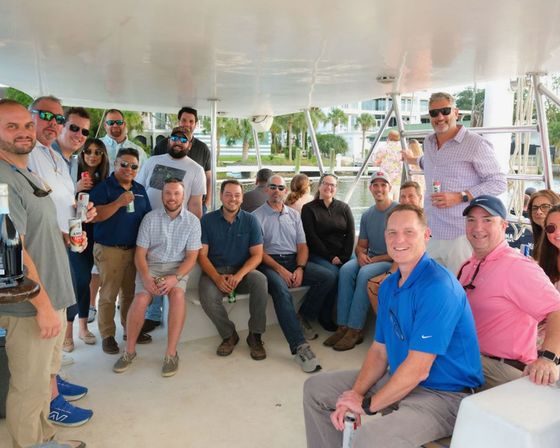 Smiling group of colleagues on a covered boat deck enjoying drinks during a sunny waterfront marina outing with palm trees and condos in the background.