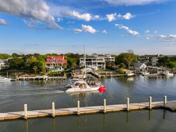 Sunlit catamaran tour boat cruising a calm marina channel past waterfront homes and private docks under a bright blue sky with scattered clouds.