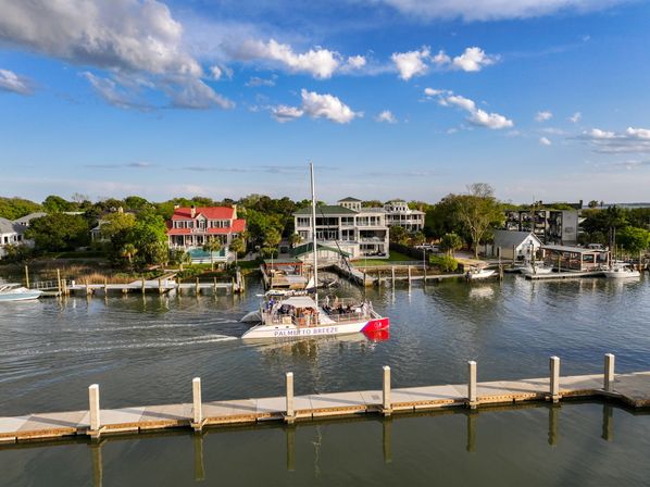 Sunlit catamaran tour boat cruising a calm marina channel past waterfront homes and private docks under a bright blue sky with scattered clouds.