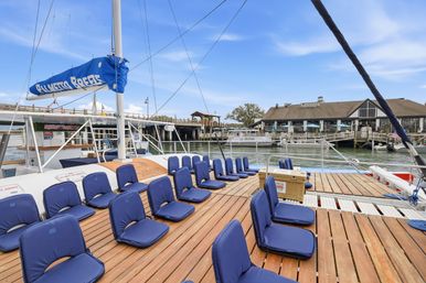Wooden sailboat deck lined with blue cushioned seats, docked at a sunny marina with waterfront restaurants, piers, and boats in the background.